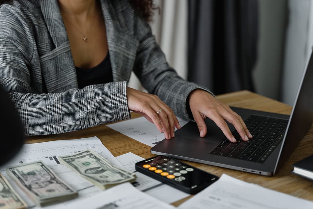 mulher mexendo no notebook ao lado de um celular com o aplicativo calculadora aberto, organizando as finanças.
